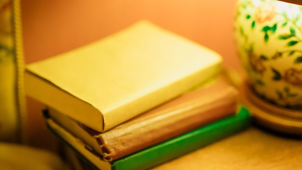 A stack of books on a bedside table under a warm lamp