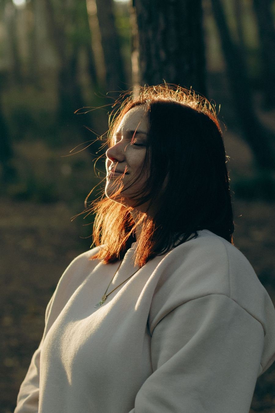 A woman with her eyes closed, smiling in warm sunlight among trees