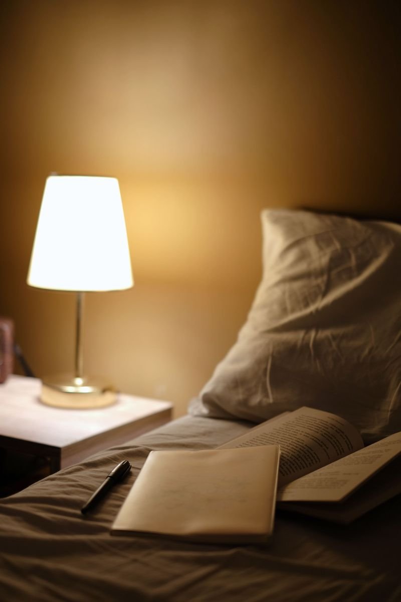 A lit bedside lamp beside a bed with an open book and a notebook